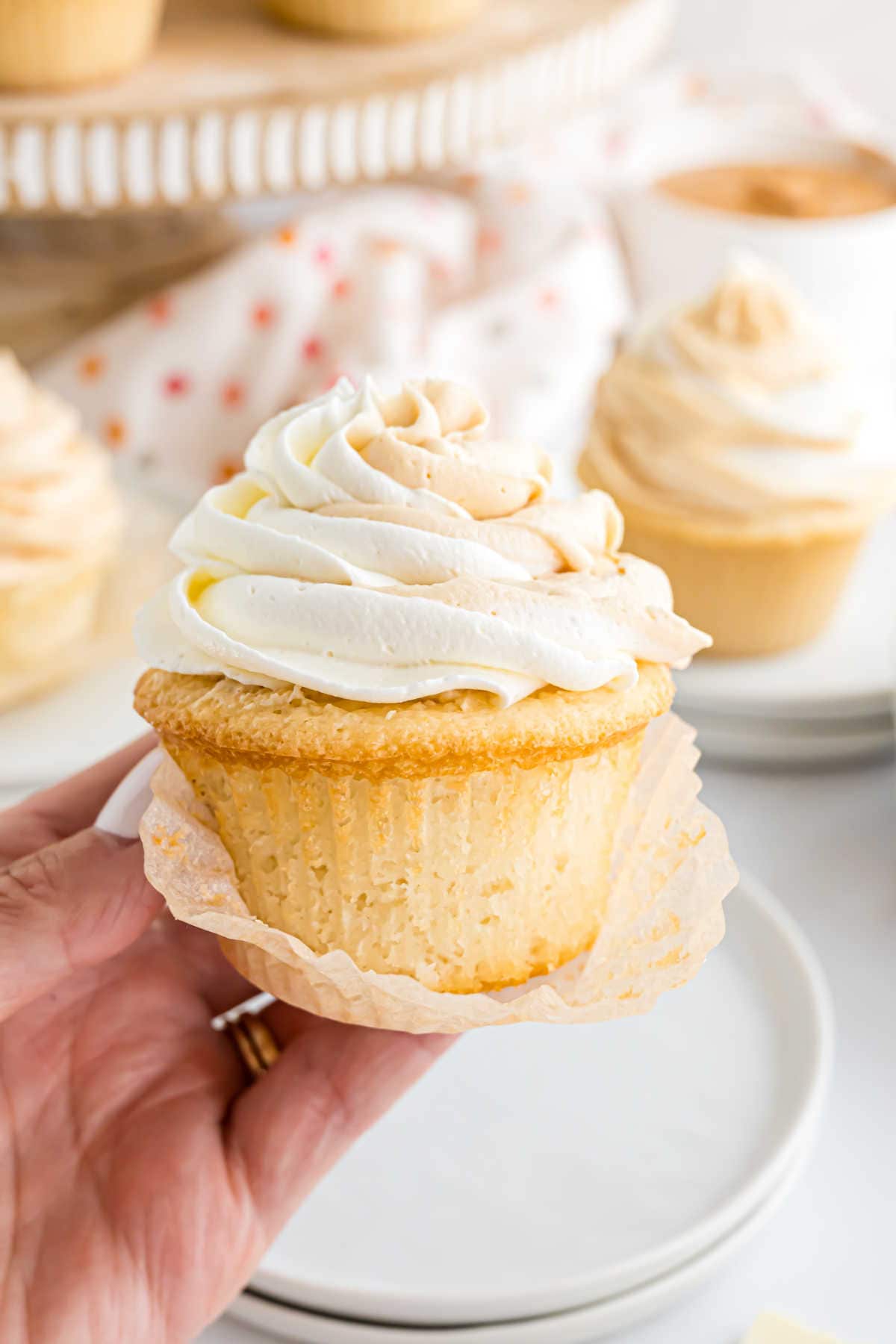 A hand holds a vanilla cupcake with swirled frosting, while white chocolate cupcakes are blurred in the background.