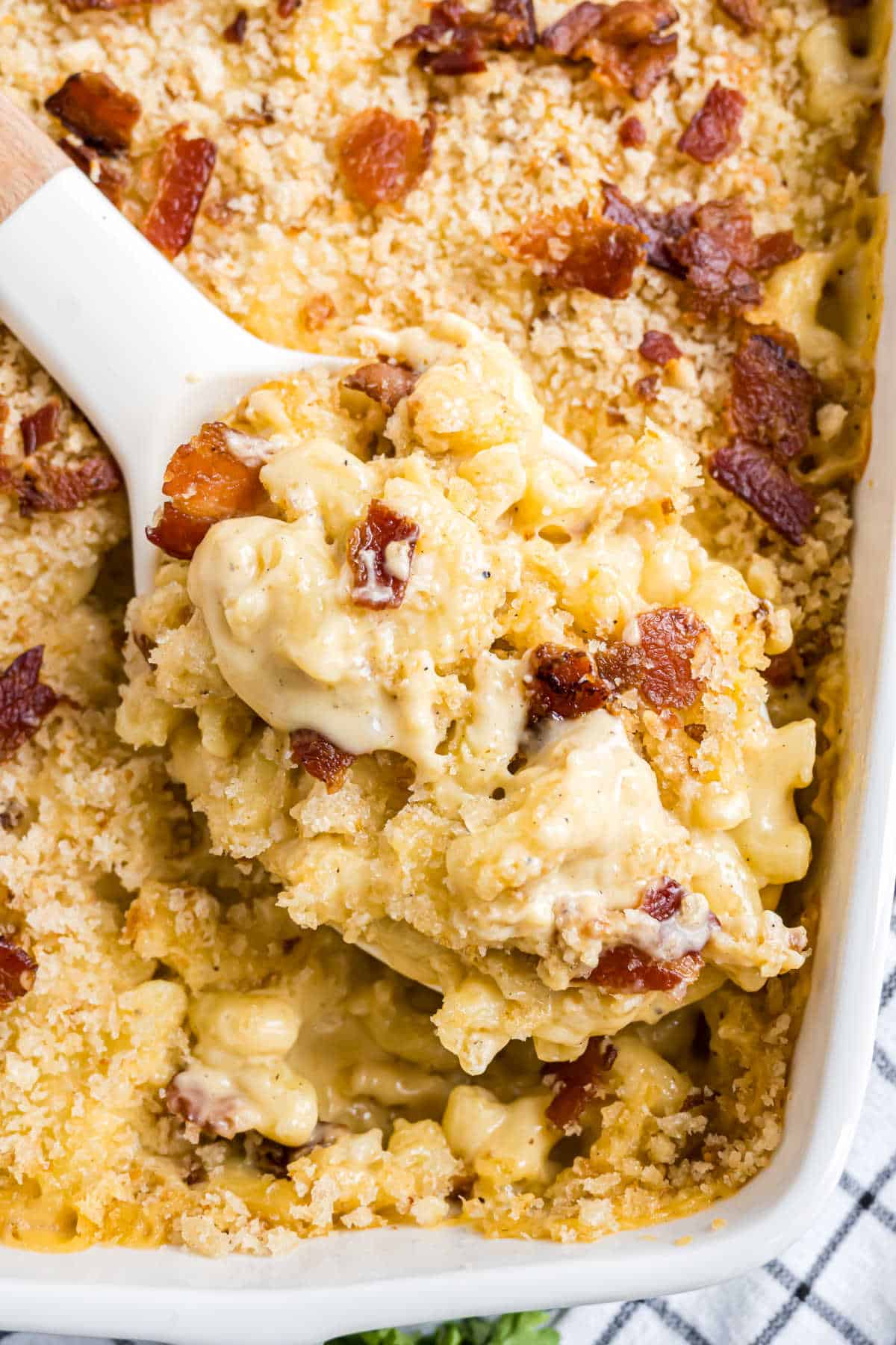 A serving spoon scoops creamy gouda bacon macaroni and cheese with crispy bacon from a baking dish.