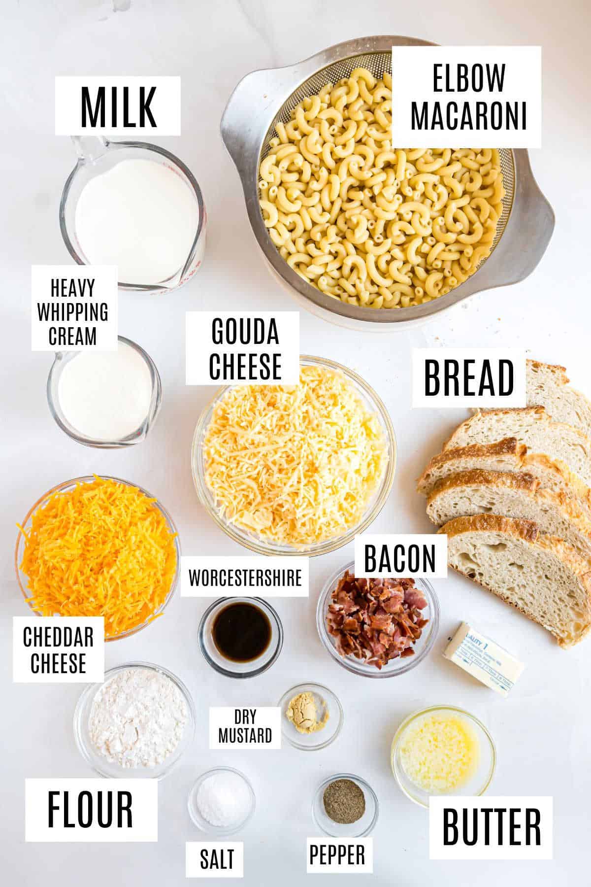 Ingredients for gouda bacon macaroni and cheese arranged on a white surface, each labeled, including pasta, cheese, and bread.