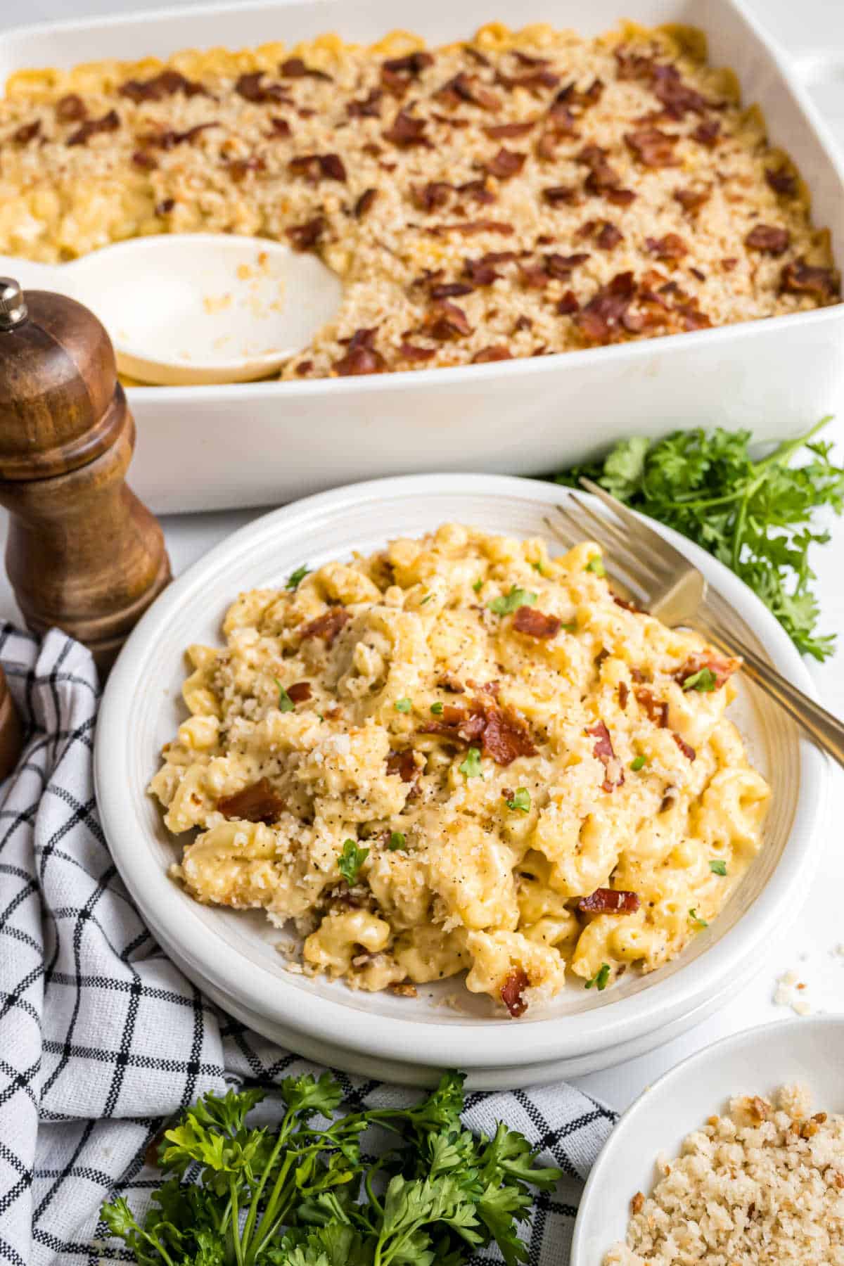 A plate of baked gouda bacon macaroni and cheese with golden breadcrumbs, next to a casserole dish and fresh parsley.