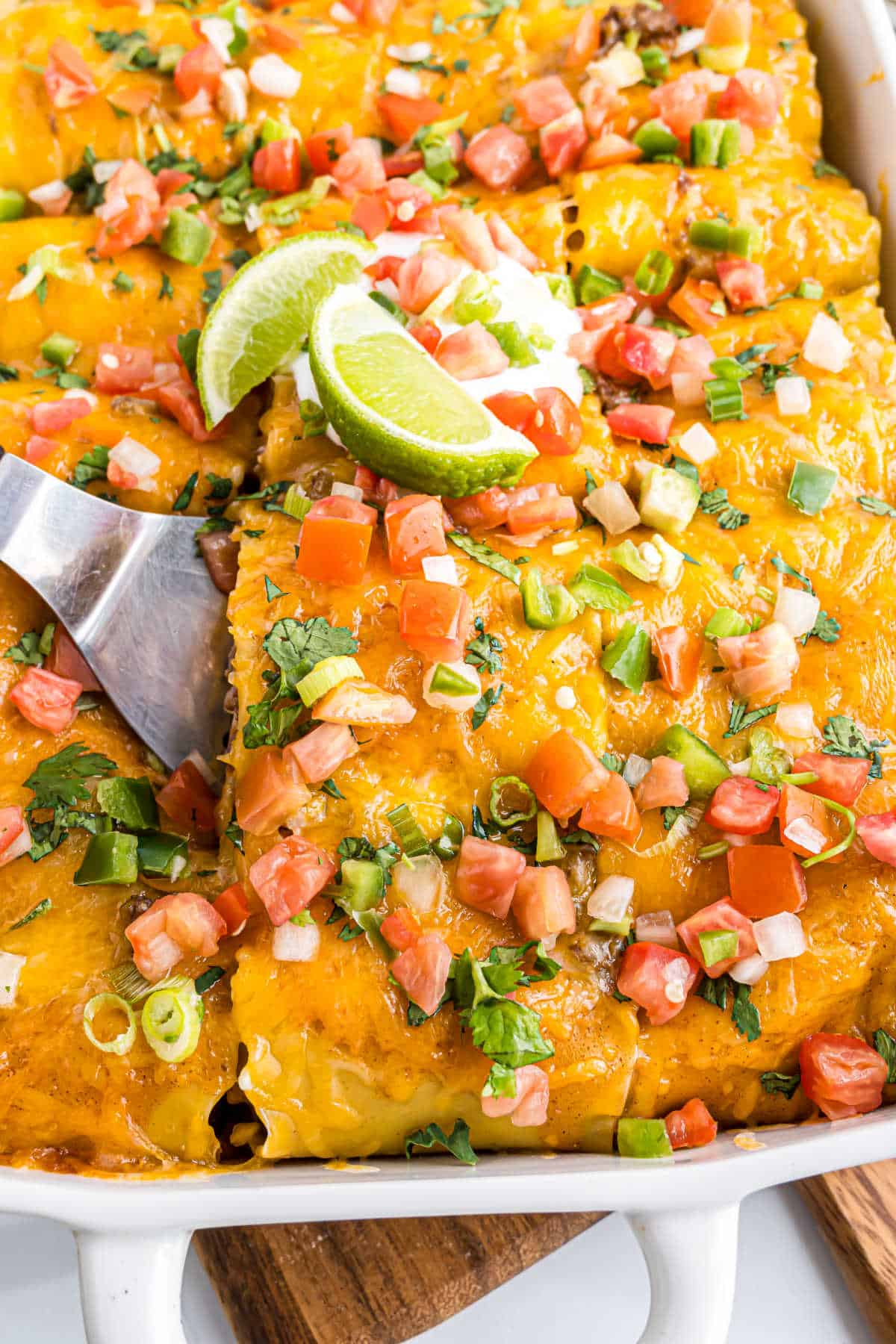 Cheesy enchilada beef rollups topped with diced tomatoes, green onions, cilantro, and lime wedges in a baking dish.