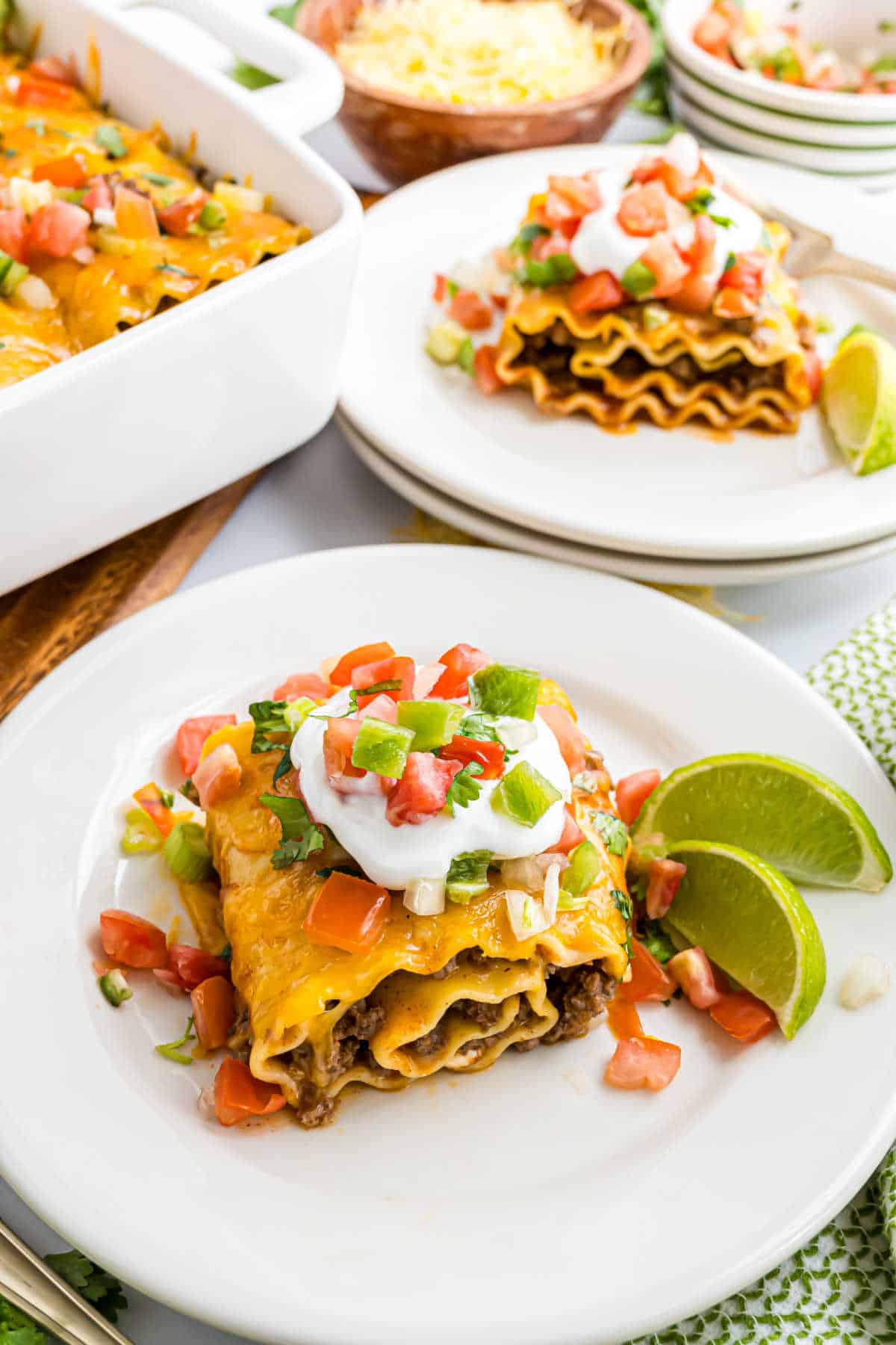 A slice of taco lasagna topped with sour cream, tomatoes, and cilantro, served with lime wedges on a white plate—perfect for fans of hearty dishes like enchilada beef rollups.