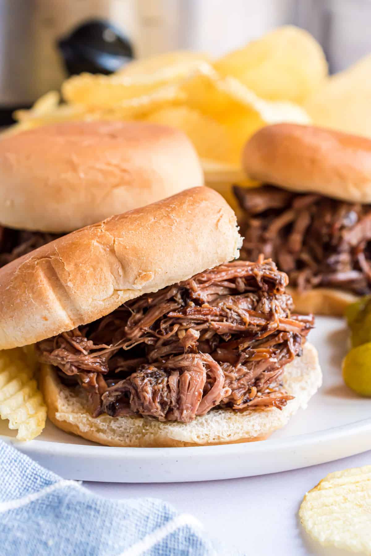Shredded beef on a bun served on a white dinner plate with potato chips.