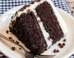 Slice of double layer chocolate cake with vanilla frosting on white plate with fork.