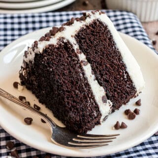 Slice of double layer chocolate cake with vanilla frosting on white plate with fork.