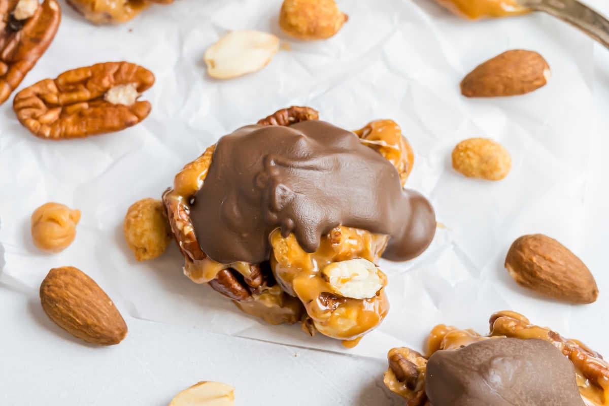 A cluster of pecans, almonds, peanuts, and caramel topped with chocolate creates irresistible caramel nut clusters, surrounded by whole nuts on parchment.