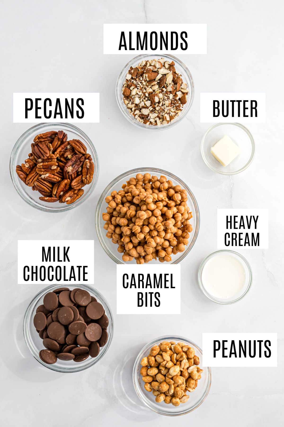 Seven bowls labeled almonds, pecans, peanuts, caramel bits, milk chocolate, butter, and heavy cream on a white surface—perfect for making caramel nut clusters.