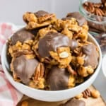A bowl of caramel nut clusters drizzled with chocolate, with more clusters and pecans in the background.