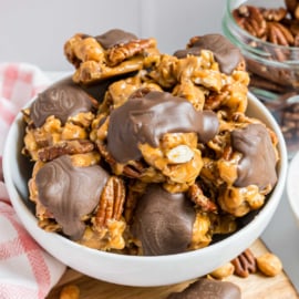 A bowl of caramel nut clusters drizzled with chocolate, with more clusters and pecans in the background.