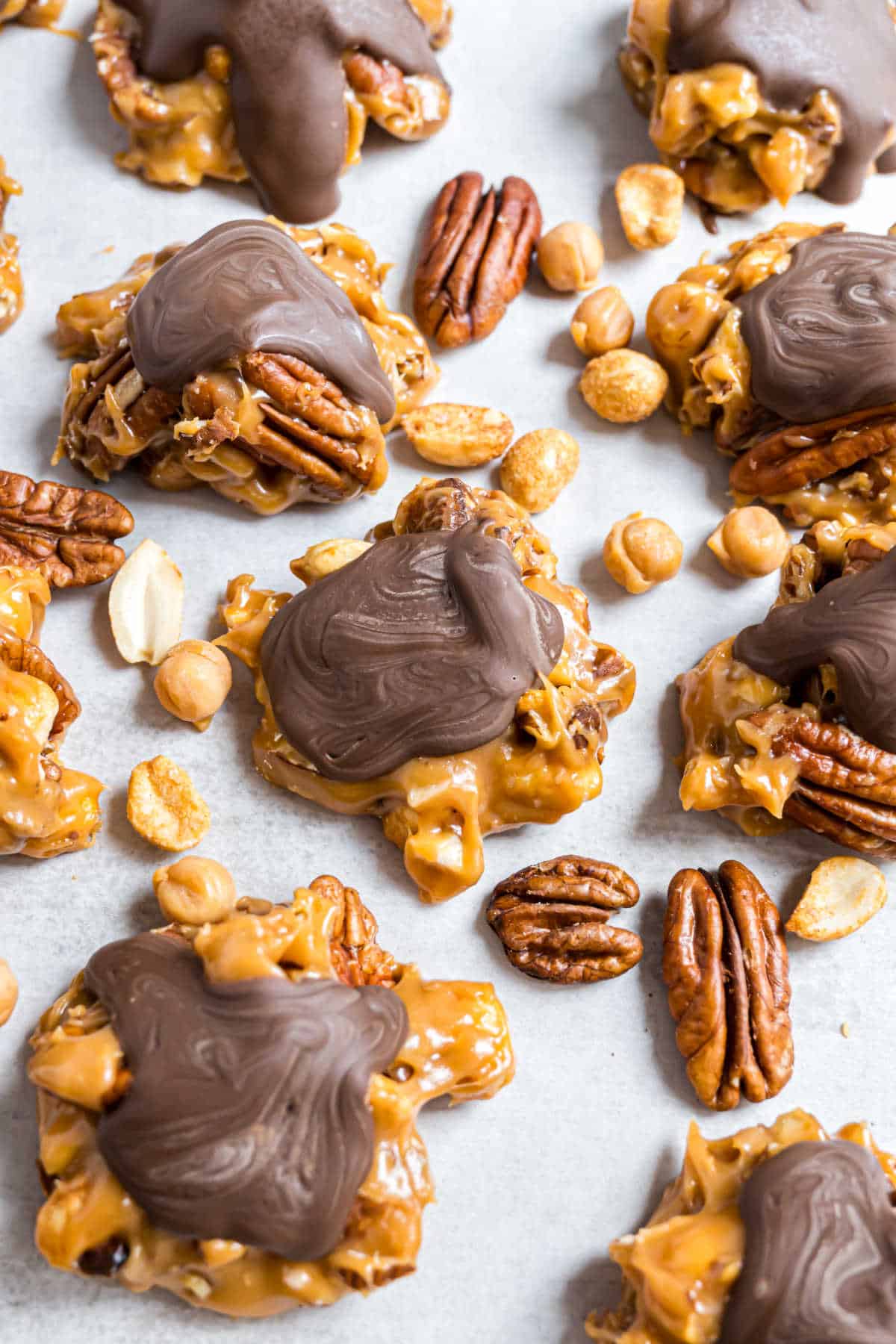 Caramel nut clusters of pecans and peanuts are coated in gooey caramel and topped with melted chocolate on a light surface.