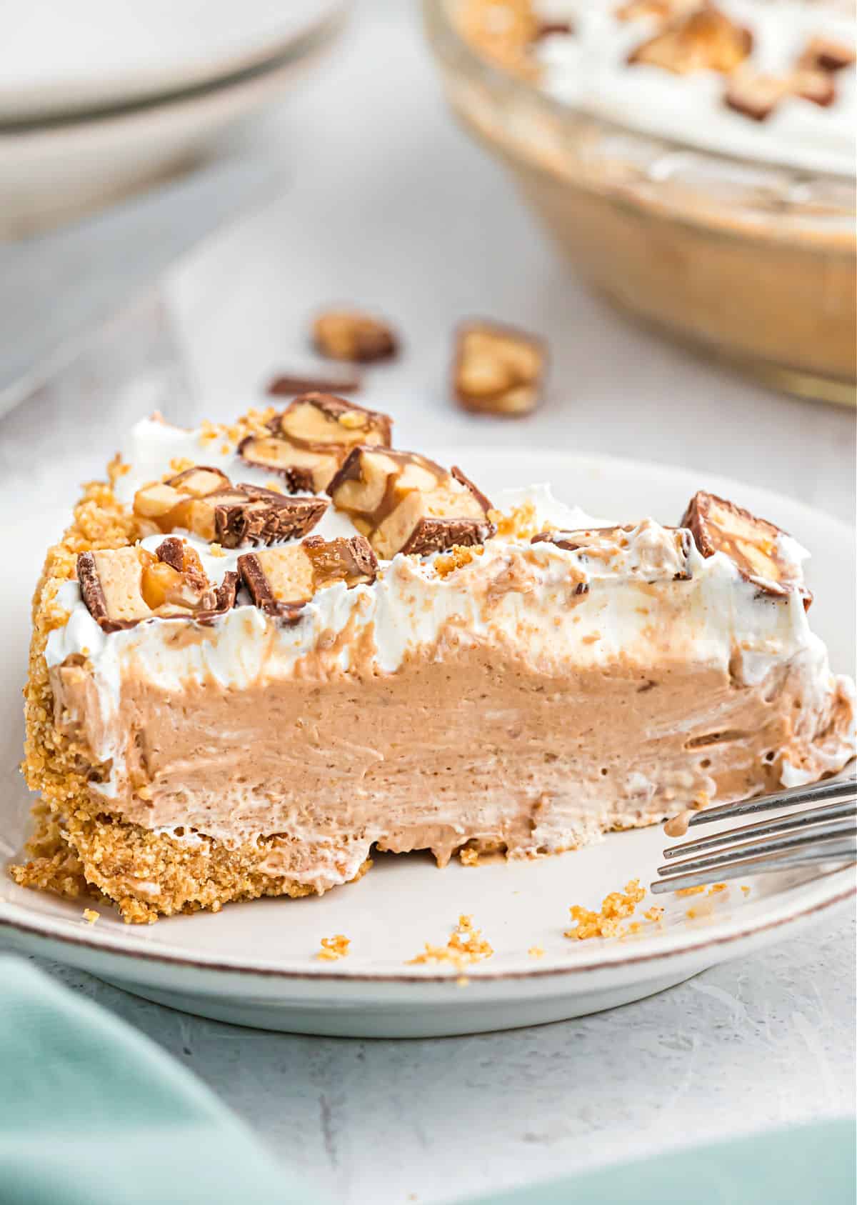 Slice of snickers pie on a white plate and topped with chopped candy bars.