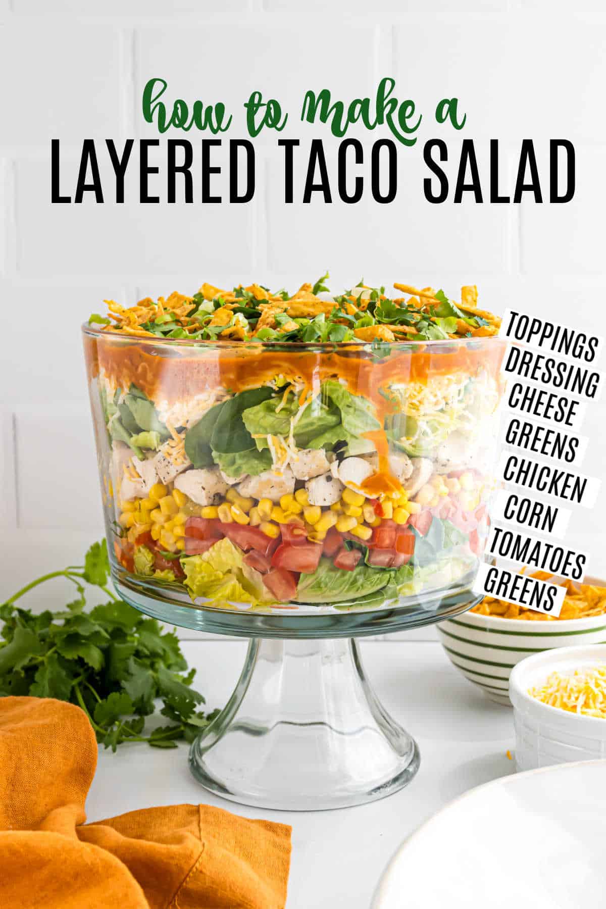 A glass trifle bowl with layered chicken taco salad sits on a counter, labeled with ingredients like chicken, corn, and greens.