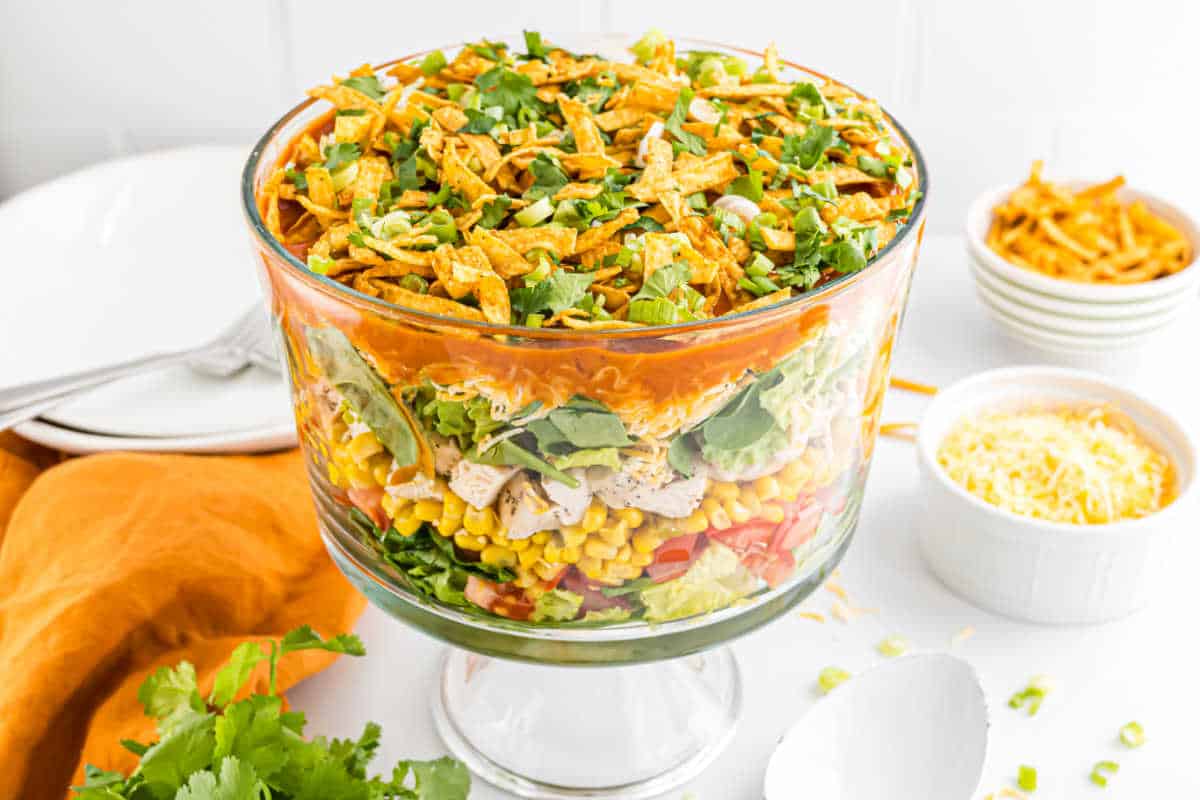 Layered chicken taco salad in a glass trifle bowl with lettuce, corn, tomatoes, cilantro, cheese, and crispy tortilla strips on top.