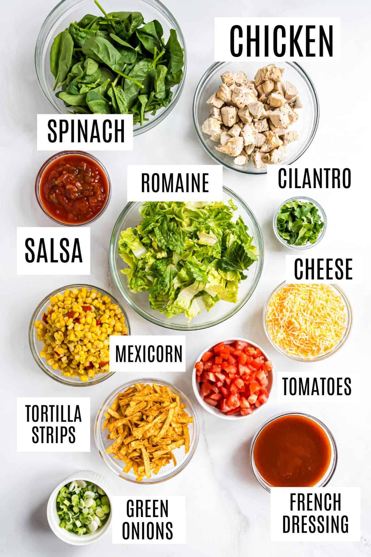 Assorted salad ingredients in bowls: chicken, greens, salsa, cheese, tomatoes, mexicorn, tortilla strips, and dressing—perfect for creating a layered chicken taco salad.