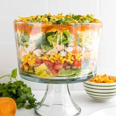 Layered chicken taco salad in a glass trifle bowl with lettuce, corn, tomatoes, cheese, and dressing, topped with fresh cilantro.