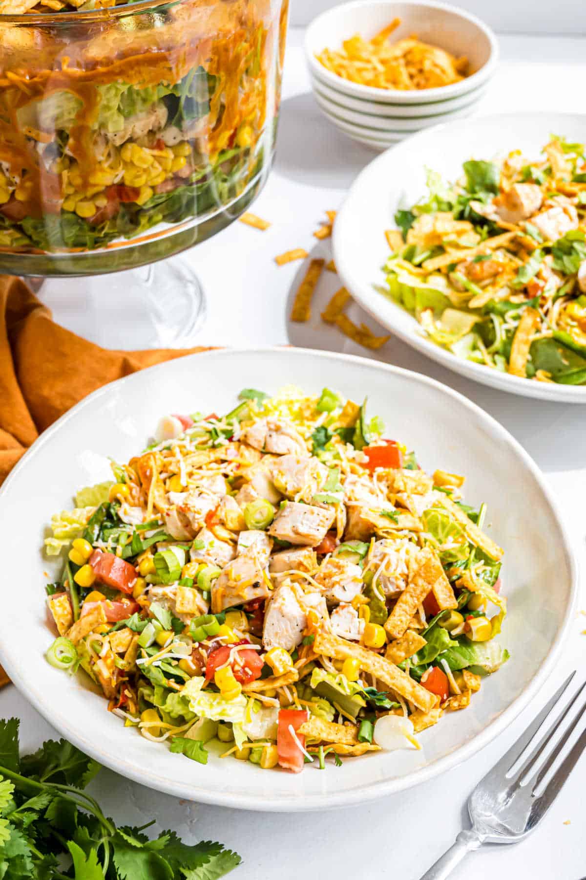A bowl of layered chicken taco salad with grilled chicken, tomatoes, corn, cheese, and tortilla strips sits on a white table.