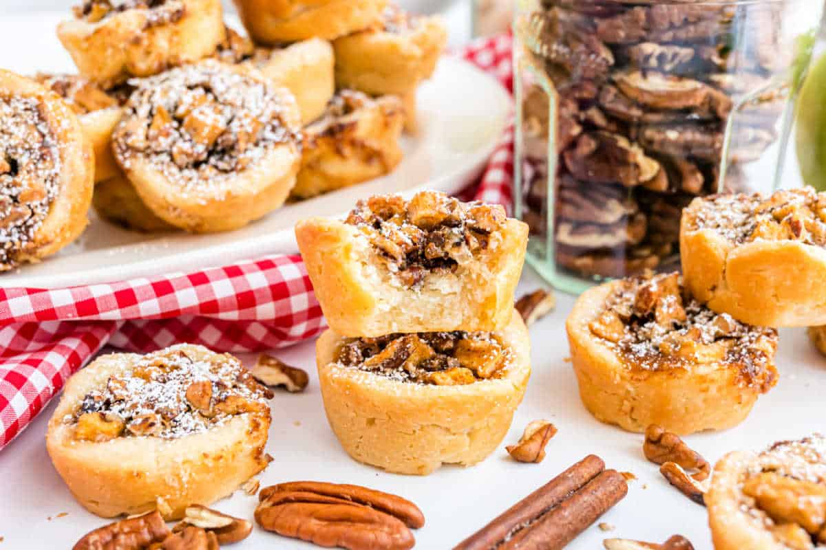 Mini pecan pies topped with powdered sugar, nestled among pecans and cinnamon sticks—these treats are the perfect complement to apple pecan cups on a festive dessert table.