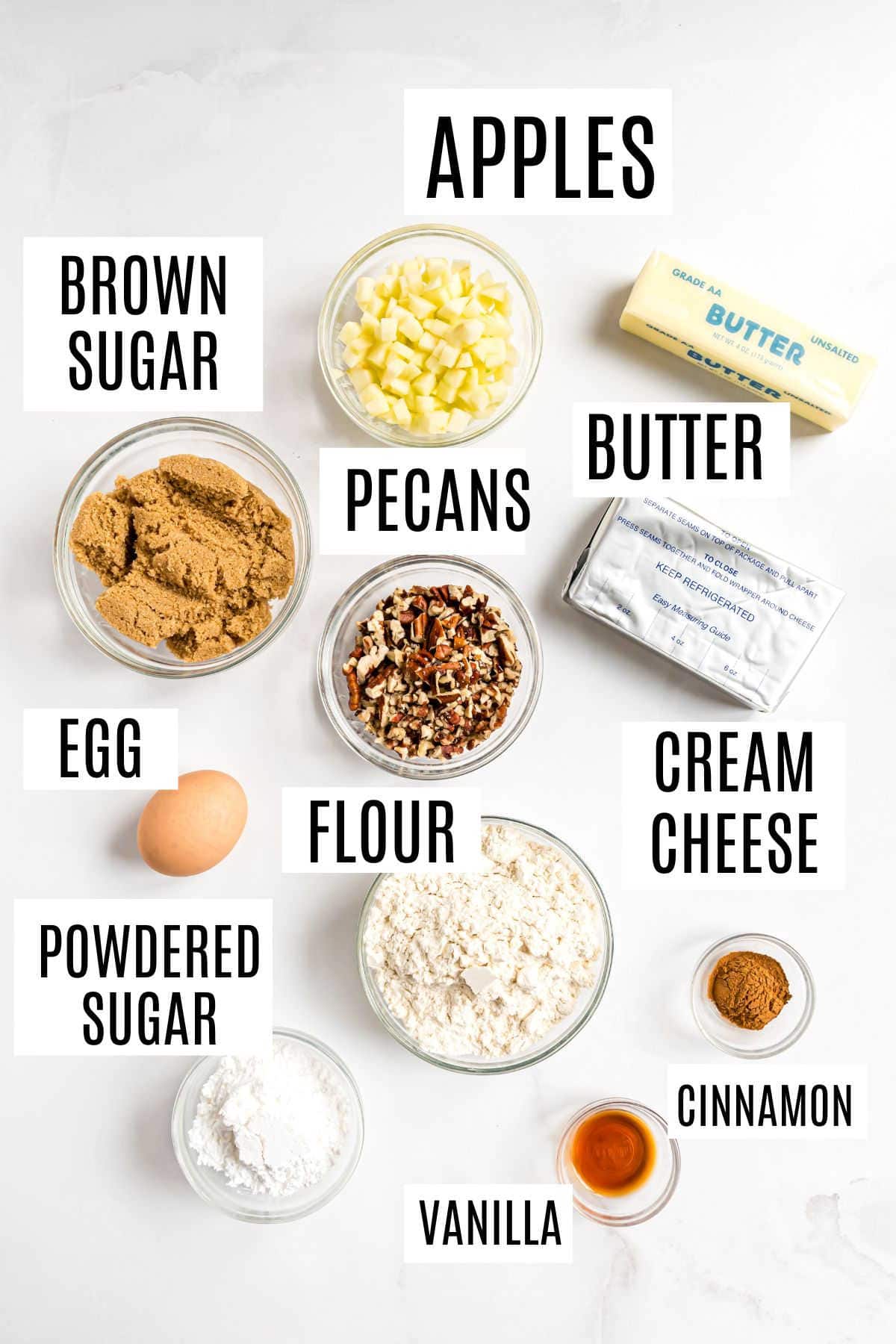 Labeled baking ingredients on a white surface, including apples, butter, flour, egg, pecans, and sugars—perfect essentials for making delicious apple pecan cups.