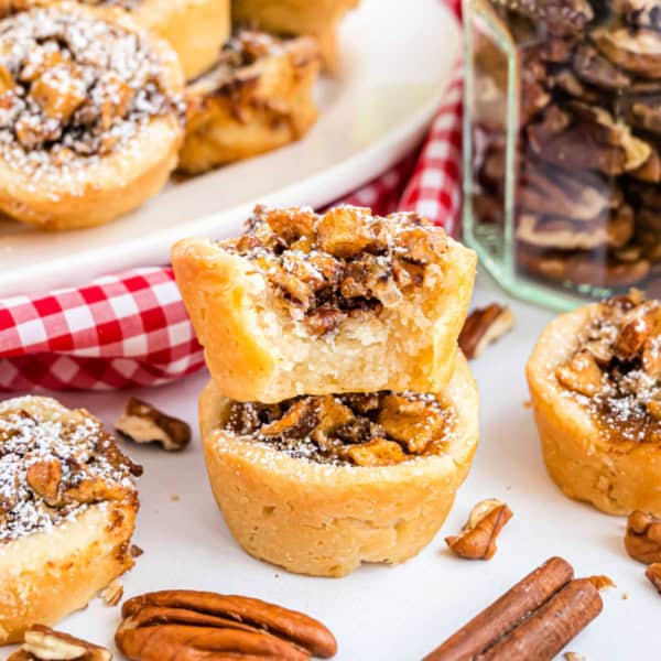 Close-up of mini pecan pies and apple pecan cups, one with a bite taken out, surrounded by pecans and cinnamon sticks.