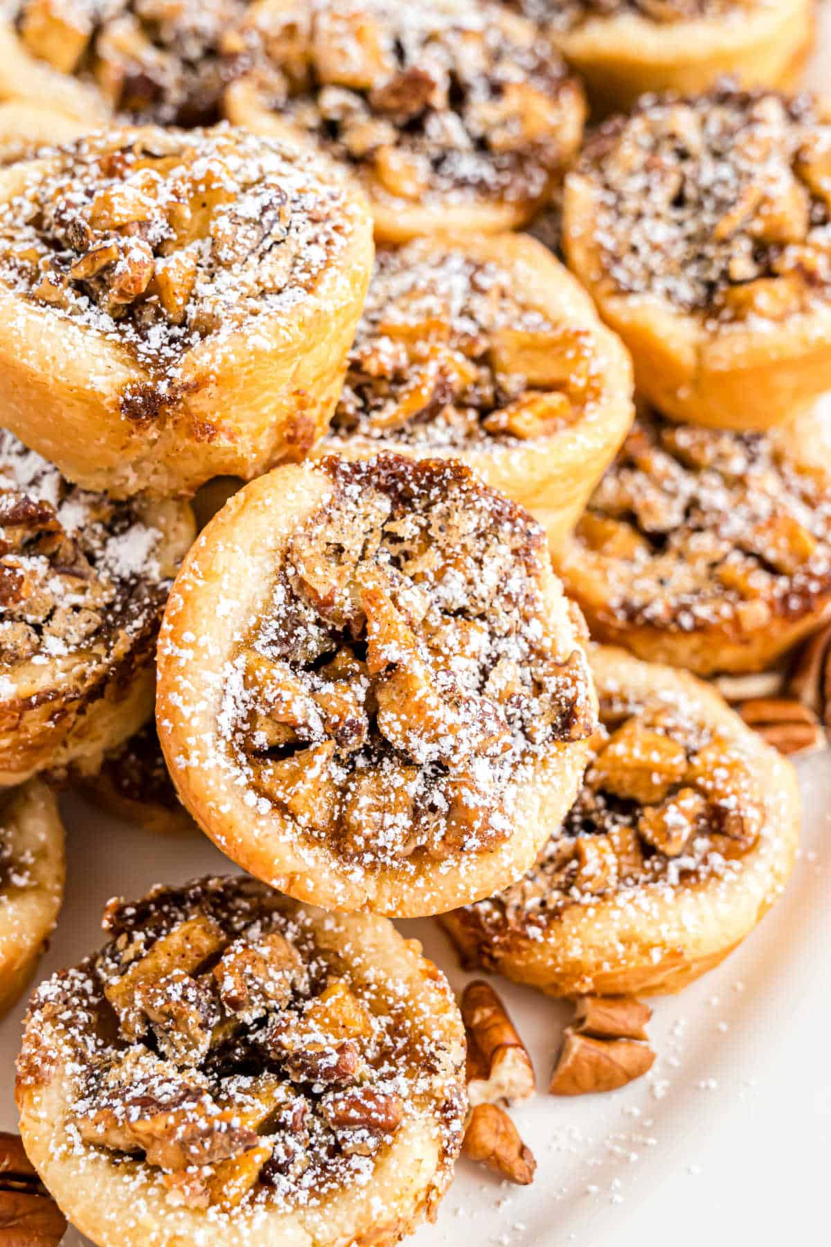 Mini apple pecan cups are stacked closely together on a white surface, each topped with a dusting of powdered sugar.