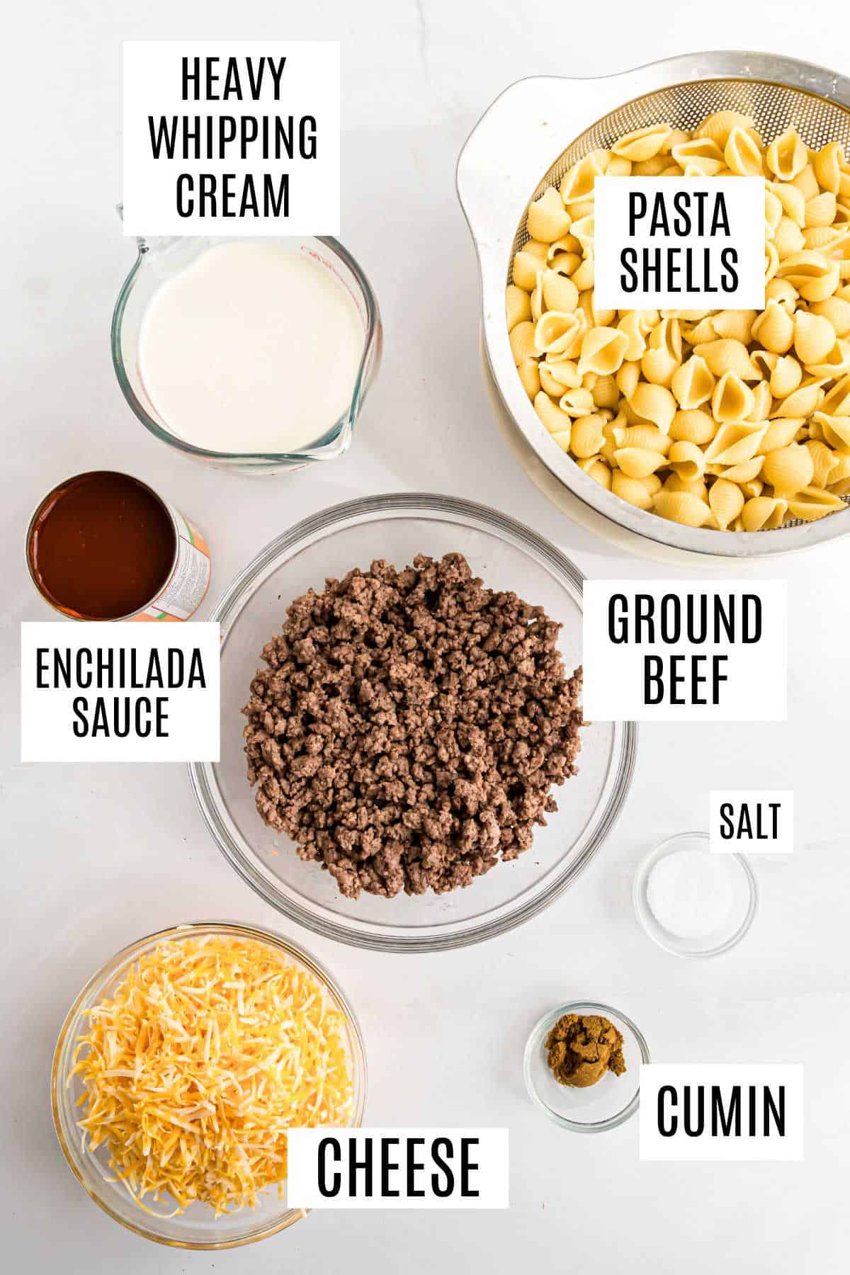 Labeled ingredients on a counter: heavy cream, pasta shells, enchilada sauce, ground beef, salt, cumin, cheese—all you need for a delicious beef enchilada mac and cheese.