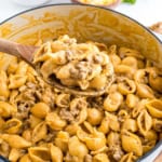 A wooden spoon scooping cheesy beef enchilada mac and cheese shells pasta with ground beef from a large pot.