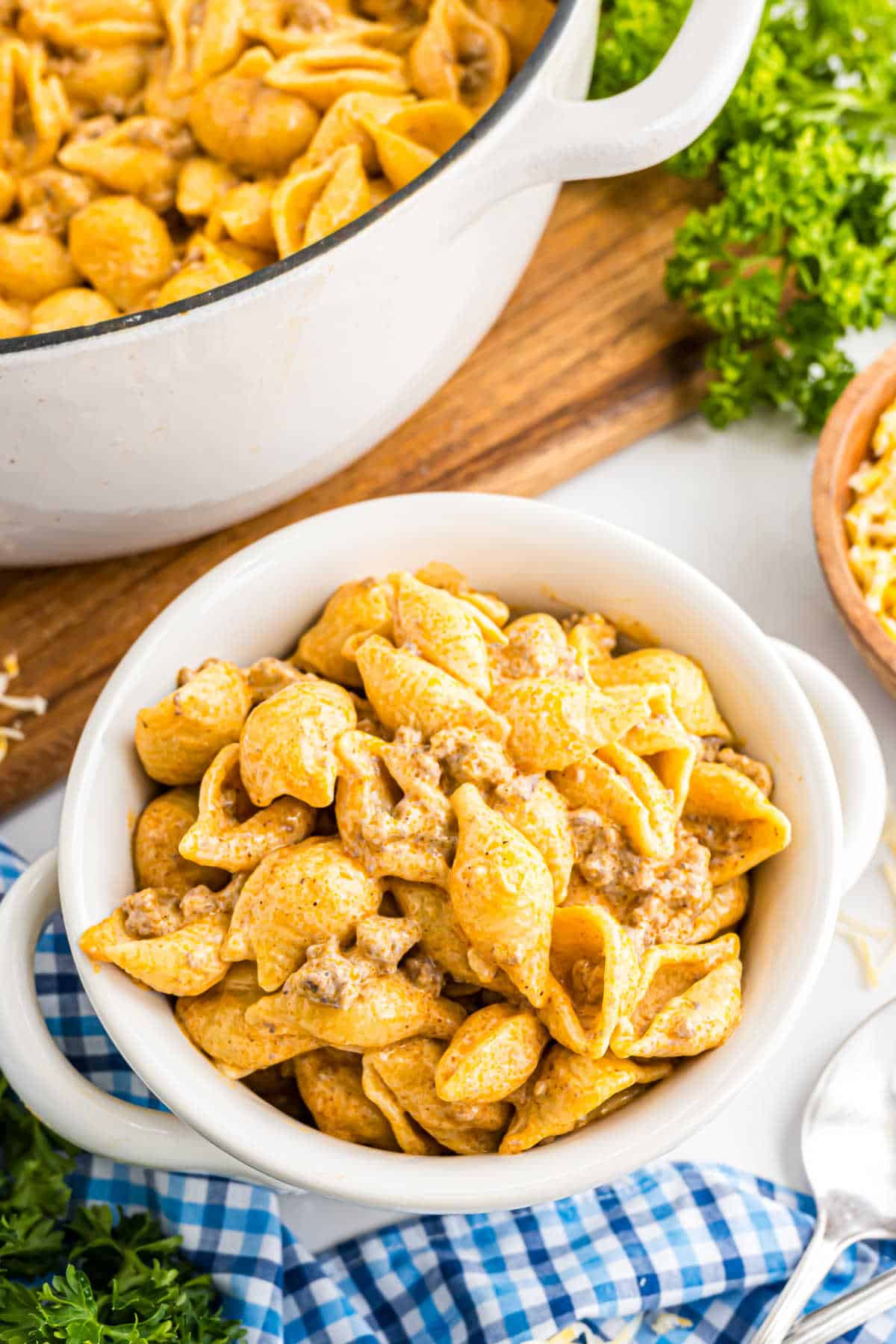 A bowl of creamy beef enchilada mac and cheese shell pasta with ground beef, next to a pot and fresh parsley on a blue checkered cloth.