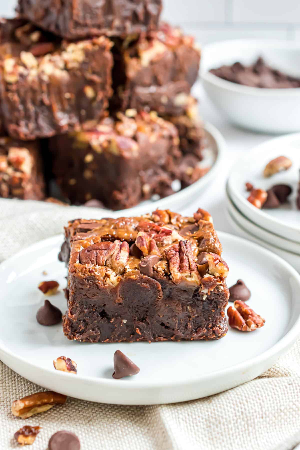 Slice of turtle brownie on a white plate with a stack of brownies in background.