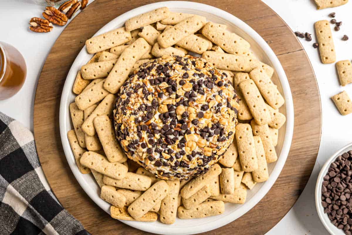 A turtle cheeseball coated with nuts and chocolate chips, surrounded by rectangular cookies on a round platter.