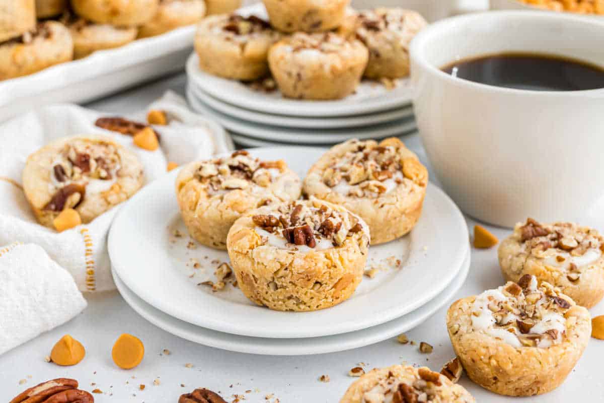 Pecan bite-sized cookies and butterscotch cookie cups sit on white plates next to a cup of coffee, with more cookies in the background.