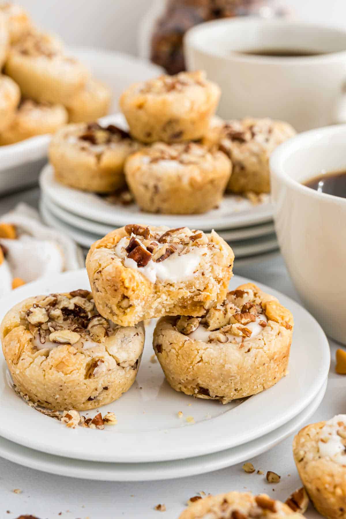 Three pecan cheesecake bites stacked on white plates, one with a creamy filling exposed, next to cups of coffee and butterscotch cookie cups.