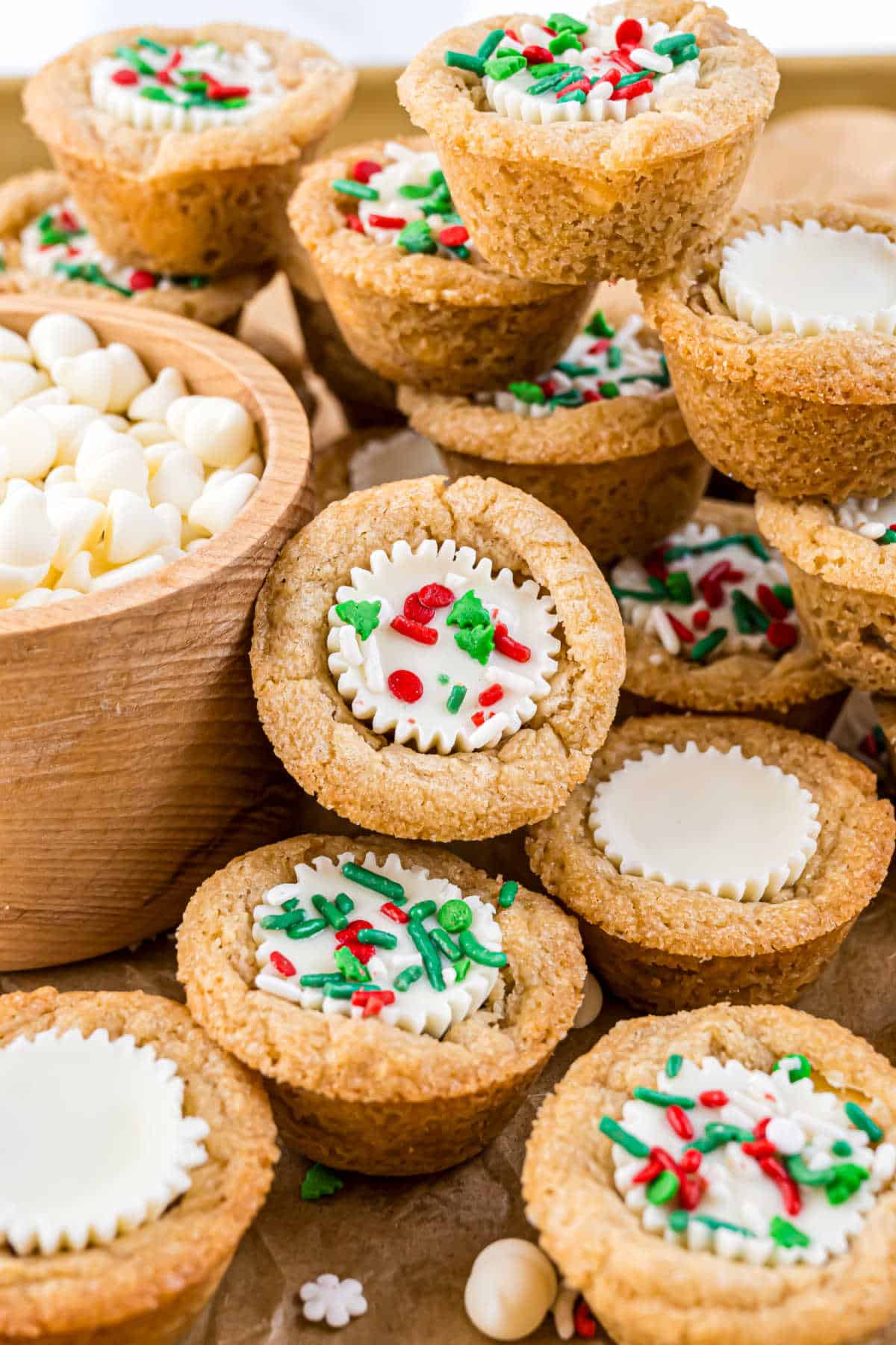 Sugar cookie cups filled with creamy white chocolate, reminiscent of white chocolate peanut butter cookies, and topped with festive red and green sprinkles, stacked on a table.