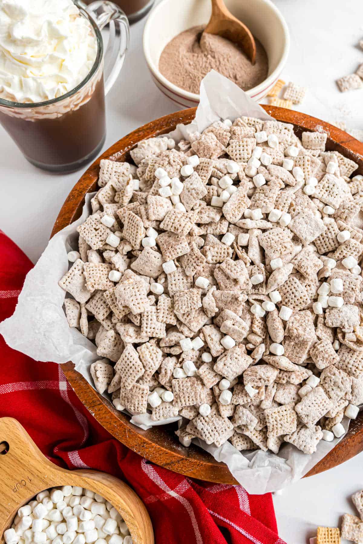 A bowl of hot cocoa muddy buddies with powdered sugar and mini marshmallows, surrounded by steaming hot cocoa and tasty toppings.