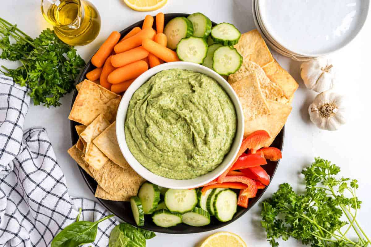 A bowl of spinach artichoke hummus surrounded by pita chips, carrots, cucumber slices, and red bell pepper strips.