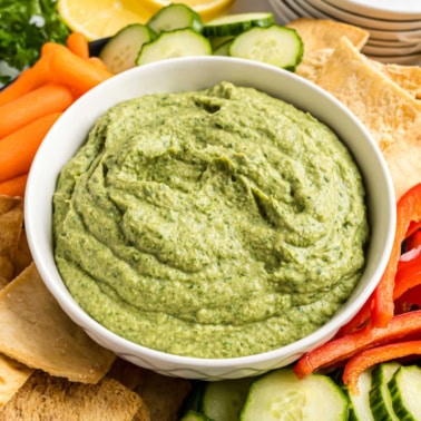 A bowl of spinach artichoke hummus surrounded by sliced vegetables, pita chips, and lemon wedges.