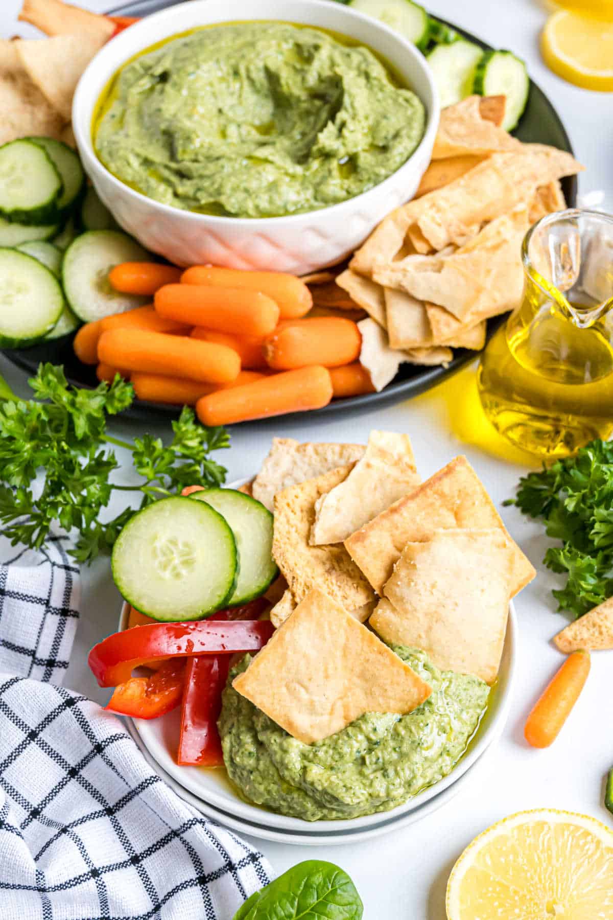 Bowl of spinach artichoke hummus served with pita chips, cucumber, carrots, and red peppers on a white table.