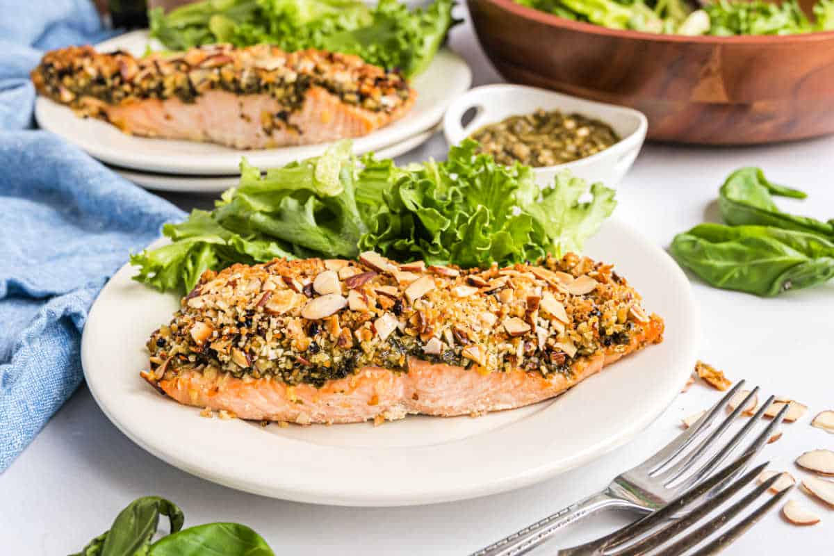 Almond pesto salmon fillet topped with chopped nuts and herbs, served with leafy greens on a white plate.
