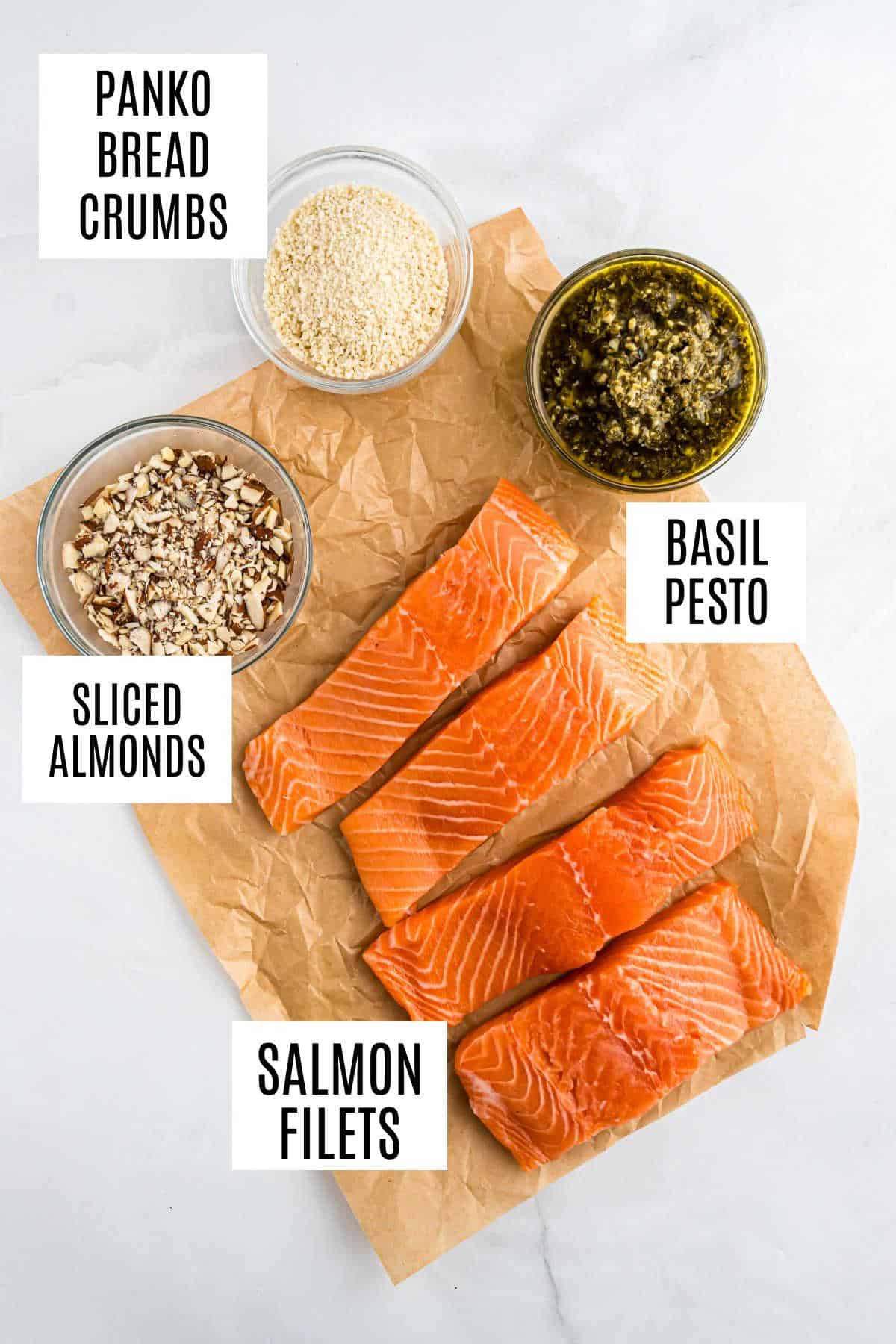 Four raw salmon fillets on parchment with bowls of panko bread crumbs, sliced almonds, and basil pesto—perfect for preparing almond pesto salmon.