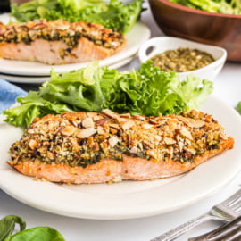 Almond pesto salmon fillet topped with herbs and sliced almonds, served with leafy greens on a white plate.