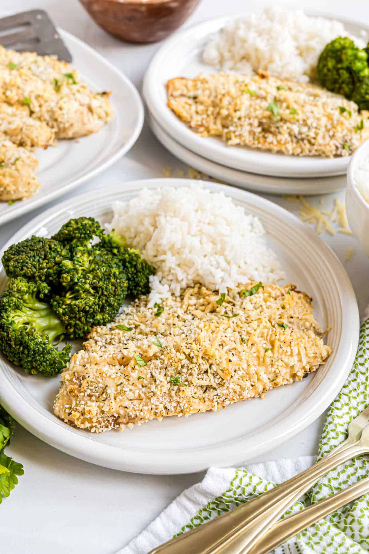 A plate with parmesan crusted tilapia, steamed broccoli, and white rice, with similar plates in the background.