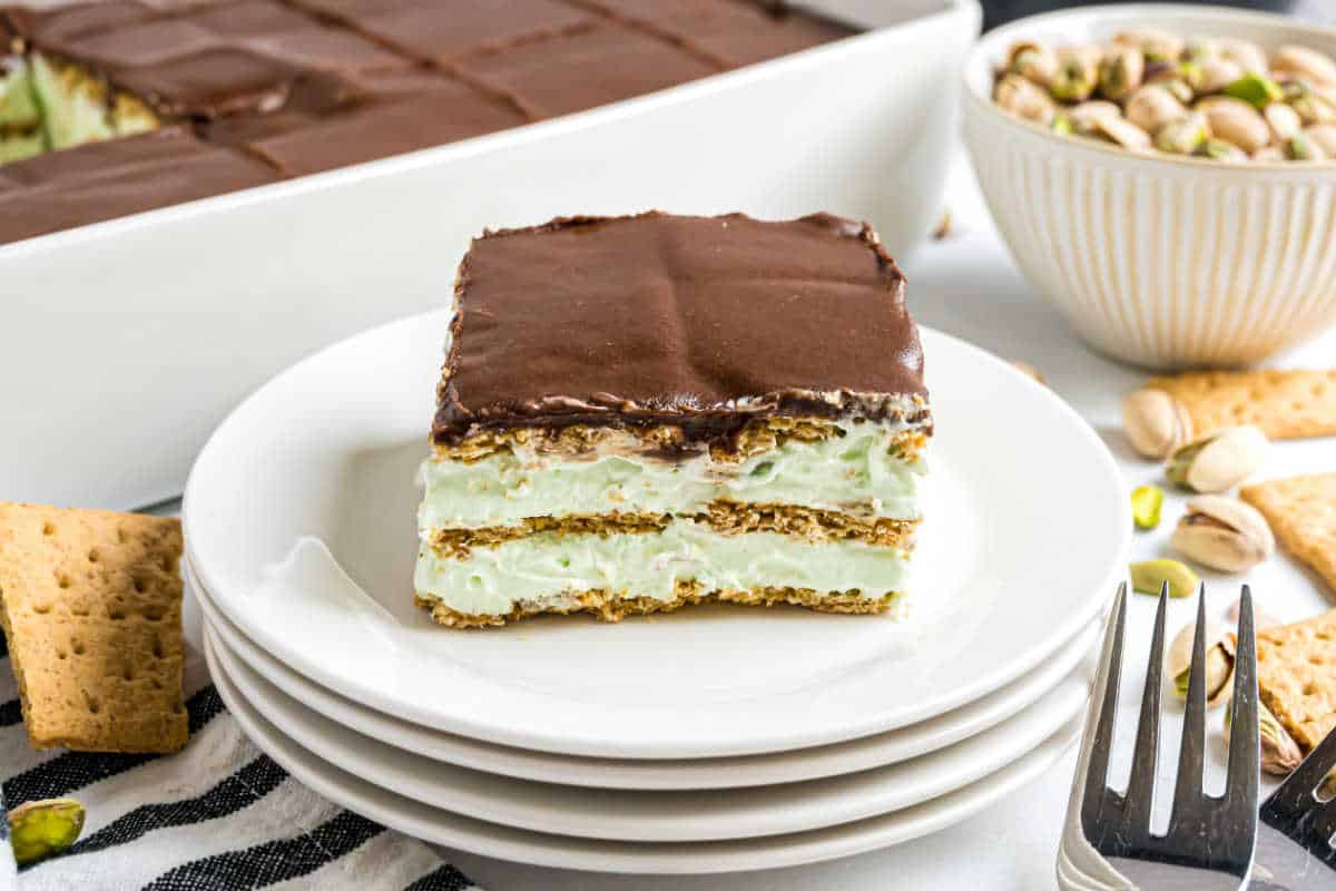 A slice of pistachio eclair cake with a rich chocolate topping sits on a white plate, surrounded by graham crackers and pistachios.