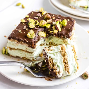 A slice of pistachio eclair cake with chocolate topping and chopped pistachios on a white plate with a fork.