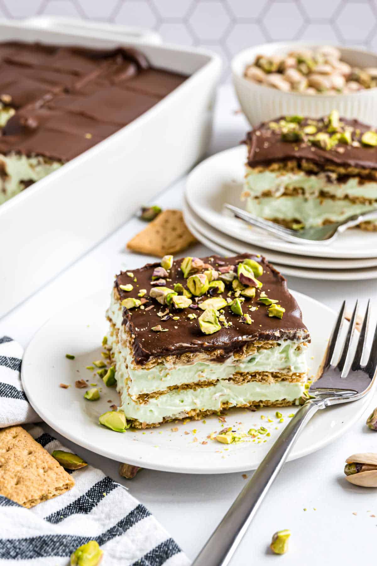 A slice of pistachio eclair cake topped with chocolate and chopped pistachios on a white plate, served with a fork.