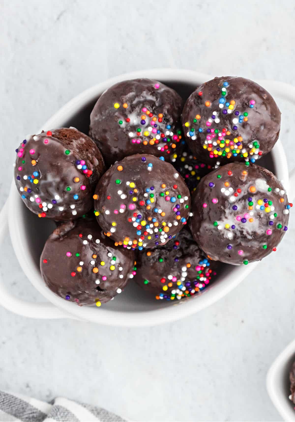 Chocolate glazed donut holes topped with colorful sprinkles in a white bowl.