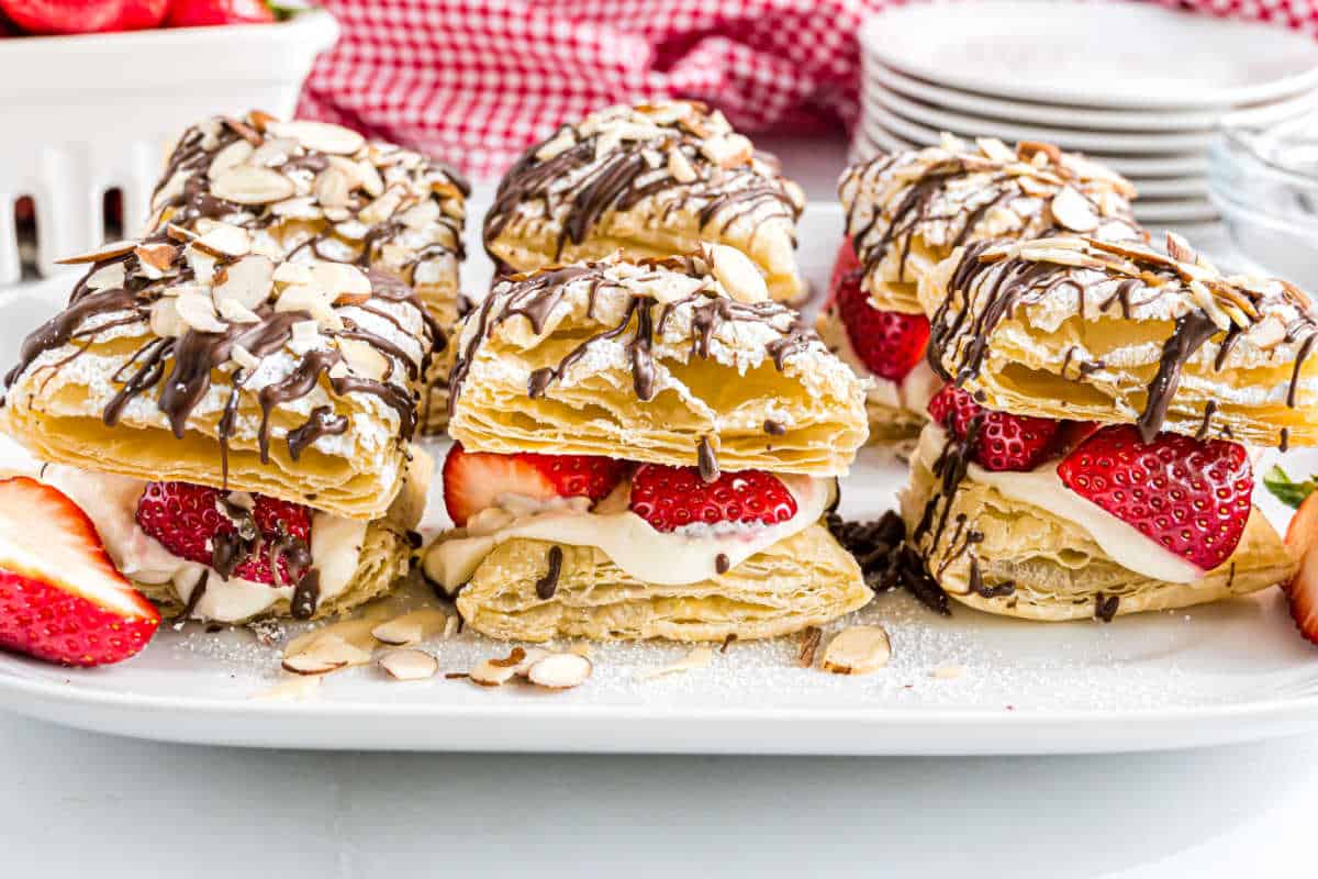 Strawberry cheesecake napoleons feature flaky pastries filled with cream and strawberries, topped with chocolate drizzle and sliced almonds on a plate.