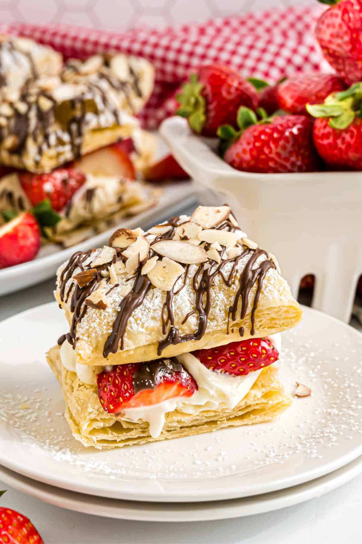 A strawberry cheesecake napoleons pastry filled with cream and strawberries, topped with chocolate drizzle and almond slices, served on a white plate.