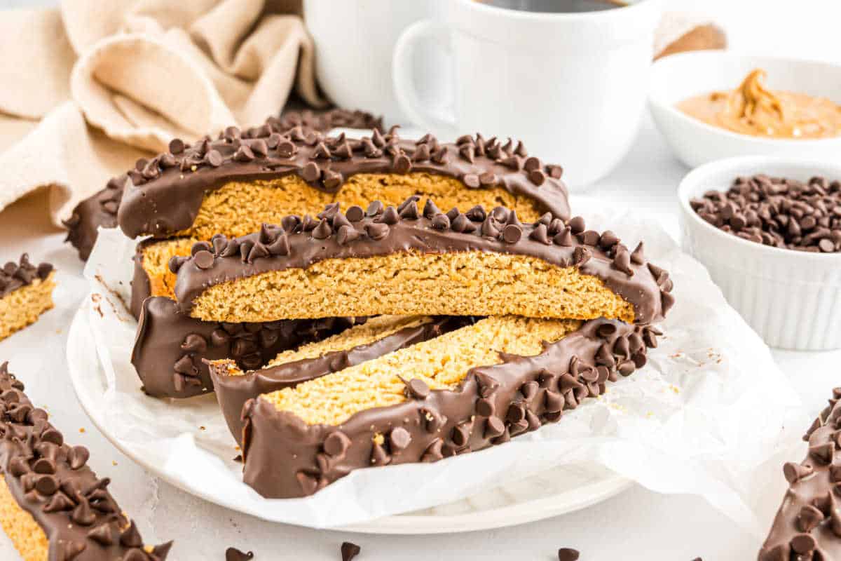 Chocolate peanut butter biscotti dipped in chocolate and topped with mini chocolate chips on a plate, with coffee and creamy peanut butter nearby.