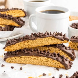 Three chocolate peanut butter biscotti dipped in chocolate are stacked on parchment beside cups of black coffee and more biscotti slices.
