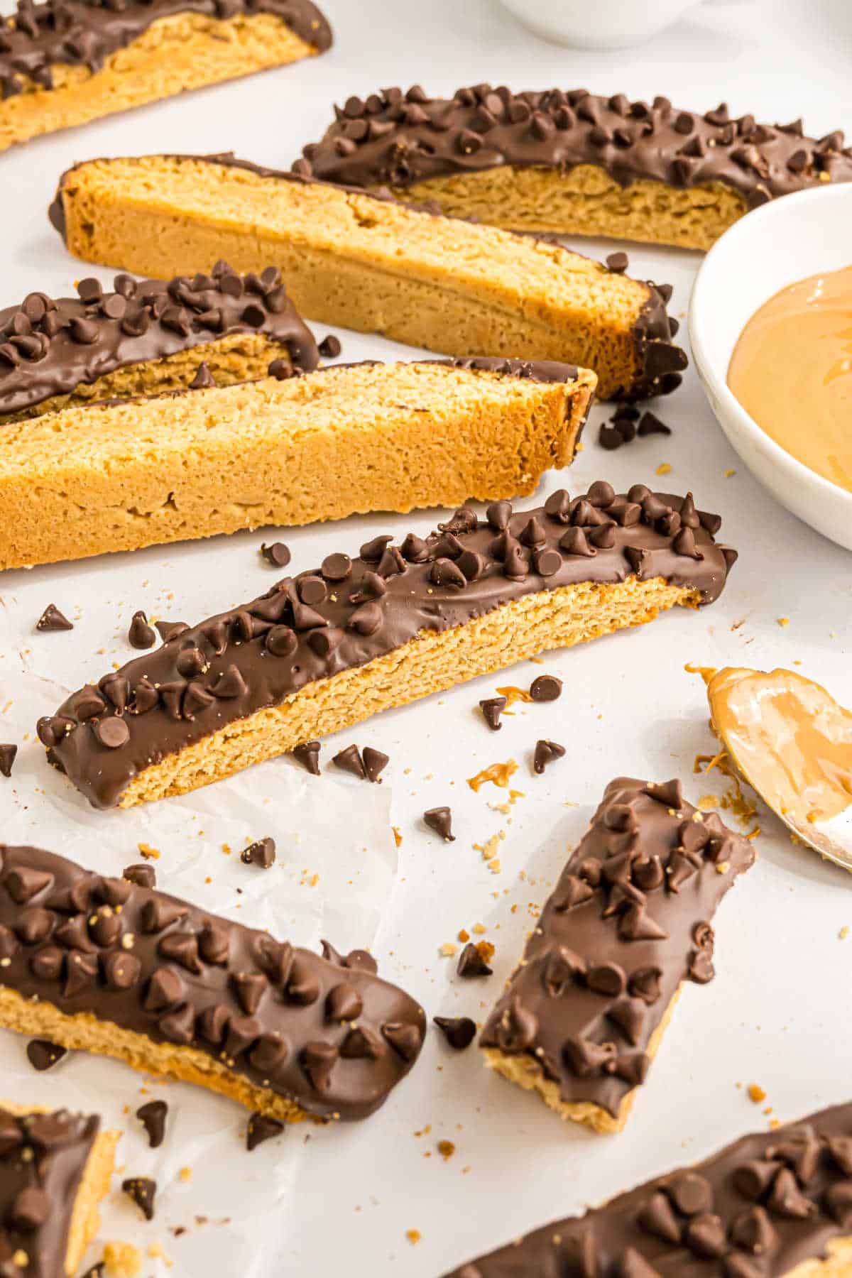Chocolate peanut butter biscotti cookies topped with chocolate and mini chocolate chips rest on parchment paper, accompanied by a bowl of creamy peanut butter.