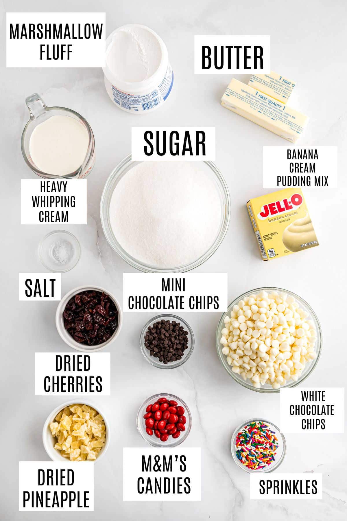 Ingredients for a delicious Banana Split Fudge dessert arranged on a white surface, each labeled, including sugar, butter, and candies.